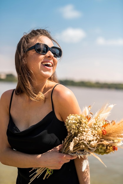 Charmante junge Dame in schwarzem Kleid und Sonnenbrille mit einem Blumenstrauß genießt einen warmen Sommertag am See