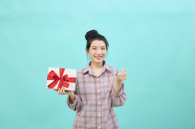 Charmante junge Frau Asiatin mit dunklen langen Haaren glücklich lächelnd in die Kamera und macht koreanische Liebeszeichen Formen Herz, mit Fingern und halten Geschenkbox auf blauem Hintergrund isoliert.