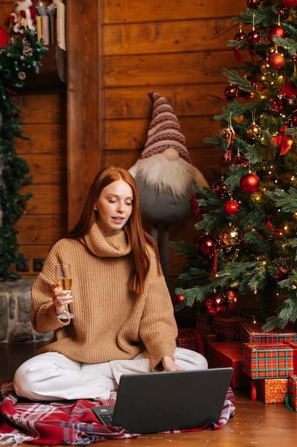 Charmante junge Frau, die auf einem Laptop spricht und Online-Glückwünsche sendet, die ein Glas mit Champagner halten