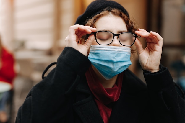 Charmante junge Frau, die auf Straße in der medizinischen Maske mit ihrer vom Atem nebligen Brille geht. Pandemiezeit und Coronavirus.