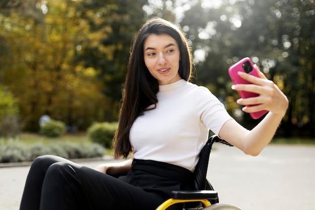 Charmante junge Frau, die ein Mibile-Telefon in der Hand hält und ein Selfie im Freien im Park macht Ein schönes Mädchen sitzt im Rollstuhl