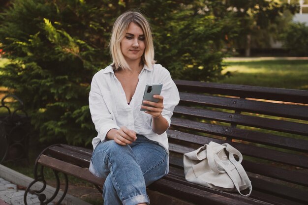 Charmante junge Frau in Freizeitkleidung sitzt auf einer Bank im Park und kommuniziert mit einem Mobiltelefon