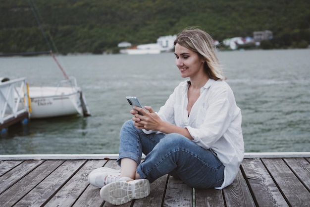 Charmante junge Frau sitzt am Pier am wunderschönen Bergsee und kommuniziert mit dem Handy