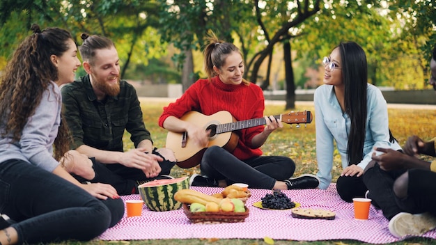 Charmante junge Frau spielt Gitarre und sitzt auf einer Decke mit Freunden auf einem Picknick Mädchen und Jungs klatschen in die Hände und hören Musik Spaß- und Naturkonzept
