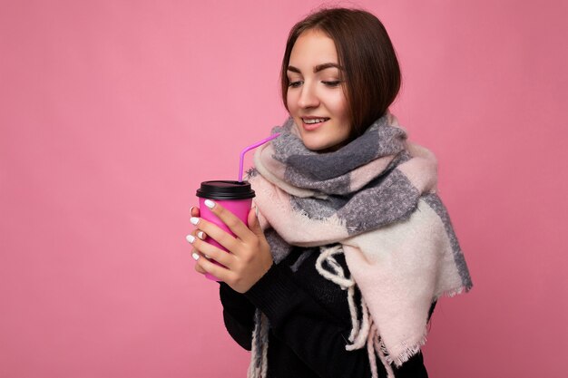 Charmante junge, glückliche brünette Frau, die stilvolle Kleidung trägt, die über einer bunten Hintergrundwand isoliert ist, die Pappbecher für Mockup-Trinken von Milchshake hält und zur Seite schaut.