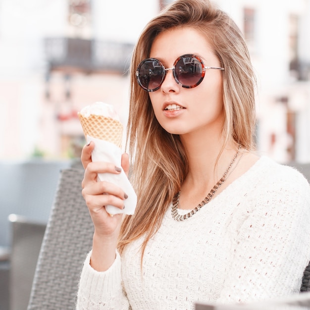 Charmante junge Hipster blonde Frau in stilvollen Sonnenbrillen in einem Vintage-Strickpullover mit süßem Eis sitzt in einem Café an einem warmen Frühlingstag. Nettes Mädchen, das ein köstliches Eis isst.