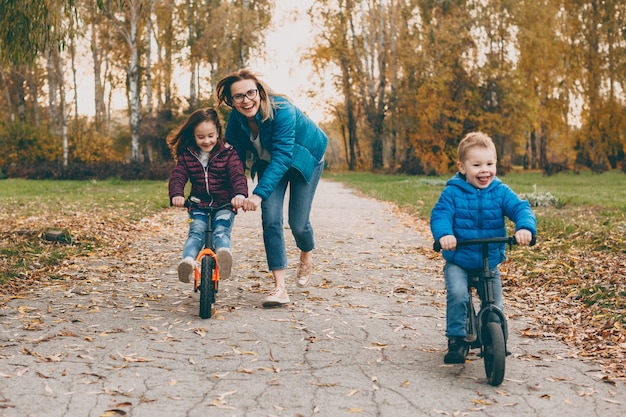 Charmante junge Mutter spielt mit ihrer Tochter und ihrem Sohn Fahrrad fahren, während der Junge geht. Mutter hilft ihrem kleinen Mädchen, Fahrrad zu fahren.
