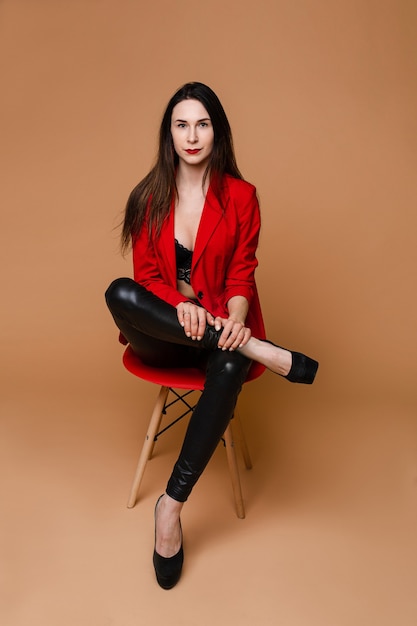 Charmante kaukasische Frau mit langen dunklen glatten Haaren in rotem und schwarzem Büroanzug, schwarze Schuhe sitzen auf rotem Stuhl