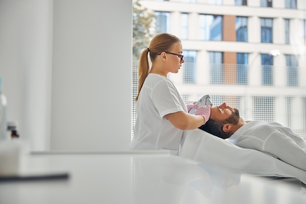 Charmante Kosmetikerin in sterilen Handschuhen, die männliche Gesichtshaut mit Lasergerät behandelt treating