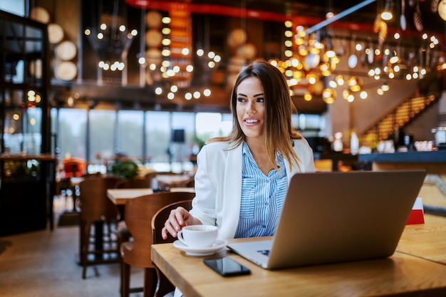 Charmante lächelnde kaukasische Brünette, die im Café sitzt und sich darauf vorbereitet, online zu arbeiten.