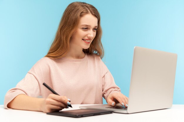 Charmante lächelnde kreative Designerin mit Grafiktablett und Tastatur-Laptop, die an professioneller digitaler Kunstausrüstung arbeitet, um Skizzen-Innenstudioaufnahme isoliert auf blauem Hintergrund zu zeichnen