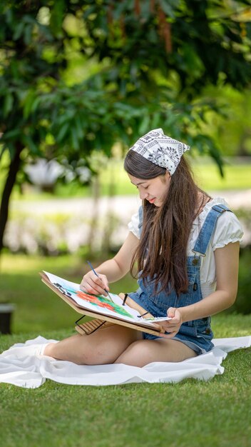 Charmante Mädchenfrau, die an sonnigen Sommertagen auf Plaid im Park sitzt und Aquarell verwendet