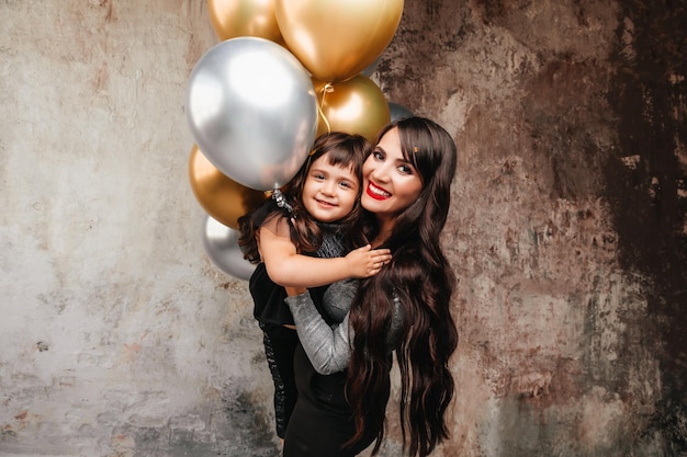 Charmante Mutter und kleine Tochter im gleichen Outfit posieren nach einer Geburtstagsfeier zusammen Porträt eines charmanten Mädchens, das ihre Tochterballons umarmt