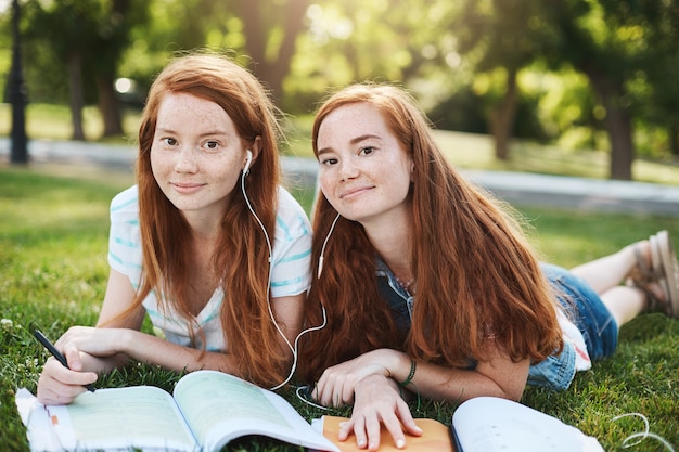 Charmante natürliche rothaarige Frauen in Sommerkleidung, die am Wochenende auf Gras liegen, Kopfhörer teilen, um gemeinsam Lieder zu hören, Schwester, die versucht, bei den Hausaufgaben zu helfen.