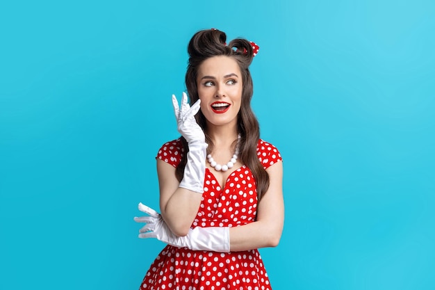 Charmante Pinup-Frau in sexy Polka-Dot-Kleid und Handschuhen mit hellem Make-up posiert auf blauem Studio