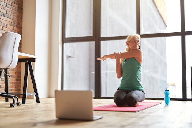 Charmante reife blonde Frau, die beiseite sitzt und auf dem Boden sitzt, während sie Yoga mit Laptop praktiziert