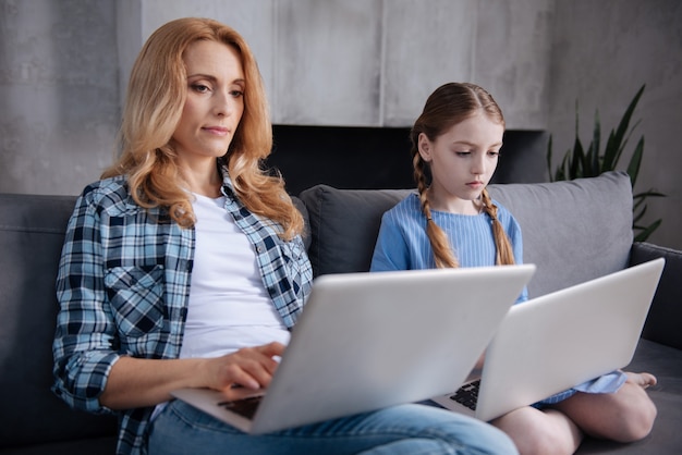 Charmante reife geschickte Mutter, die zu Hause sitzt und Laptop benutzt, während sie mit Tochter im Internet surft und tippt