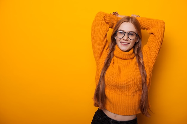 Charmante rothaarige Studentin mit roten Haaren und Brille streckt ihre Hände, während sie in einem orangefarbenen Pullover auf gelbem Grund posiert