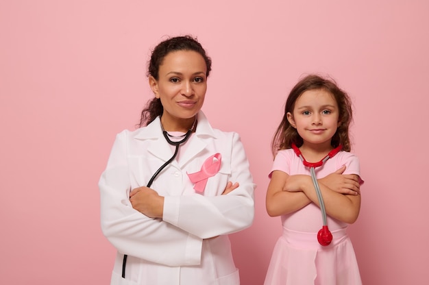 Charmante Ärztin in weißem medizinischem Kittel steht neben einem süßen kleinen Mädchen in Rosa, beide mit verschränkten Armen auf der Brust und mit einem rosa Band, Symbol des Brustkrebs-Bewusstseinstages. Medizinisches Konzept