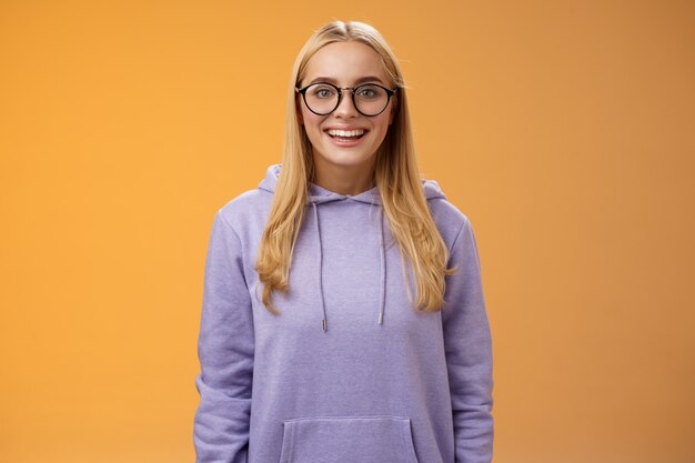 Charmante Studentin Geek lächelnd energiegeladen wollen teilnehmen Universitätsveranstaltung grinsend aussehen aufgeregt tragen Brille lila Hoodie stehen amüsiert orange Hintergrund Spaß haben. Speicherplatz kopieren