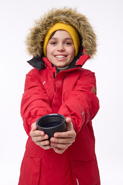 Charmanter fröhlicher Junge im Schulalter in hellen, warmen Kleidern lächelt zahnig, schaut in die Kamera und wärmt seine Hände auf einer Tasse mit einem heißen Teegetränk. Winterurlaub-Reisekonzept mit Kopien-Werberaum