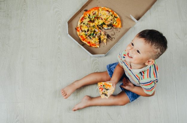 Charmanter Junge, der auf dem Boden sitzt und Pizza von oben isst?