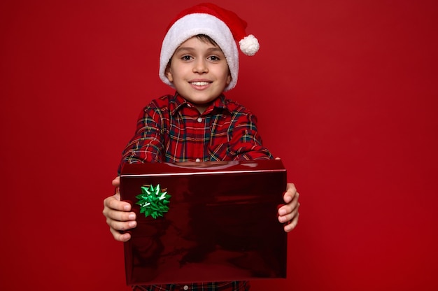 Charmanter Junge in Weihnachtsmütze und rotem kariertem Hemd hält ein Weihnachtsgeschenk in rotem Glitzerpapier mit glänzendem grünem Bogen in seinen ausgestreckten Händen und zeigt es der Kamera. Platz für Anzeige kopieren