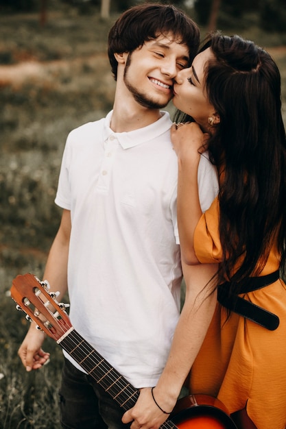 Charmanter junger Mann, der eine Gitarre hält und lacht, während seine Freundin draußen küsst, während sie in ihrer Urlaubszeit reist.