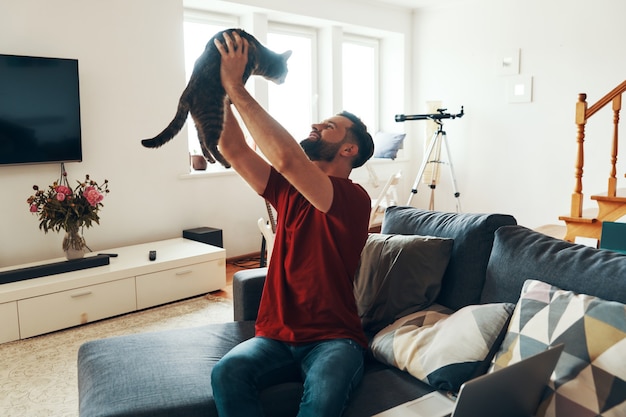 Charmanter junger Mann in Freizeitkleidung, der sich mit Hauskatze verbindet und lächelt, während er Zeit drinnen verbringt