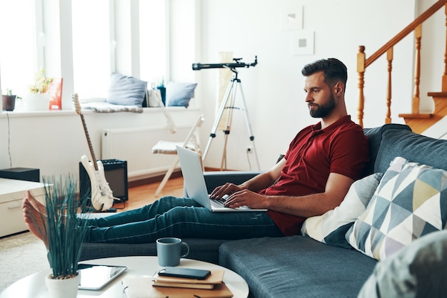 Charmanter junger Mann in Freizeitkleidung mit Laptop beim Sitzen auf dem Sofa