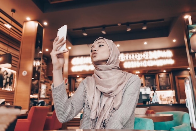 Charmantes arabisches Mädchen im Hijab macht Selfie auf ihrem Smartphone, sitzt in einem Café