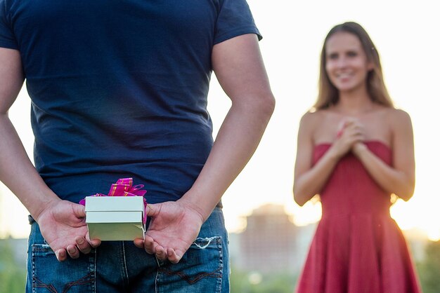 Charmantes, attraktives, schönes blondes mädchen mit langen haaren, das im abendcocktailkleid verliebt ist und auf eine geschenkbox von liebhaberjungen im sommerpark mit grasgrünhintergrund wartet. valentinstag-konzept