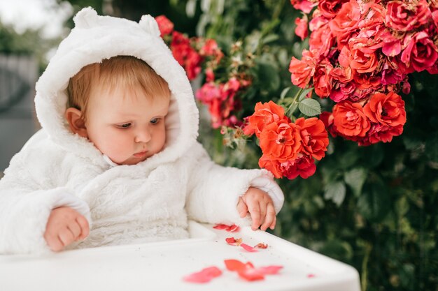 Charmantes Baby, das im Hochstuhl draußen in den Büschen mit Blumen sitzt