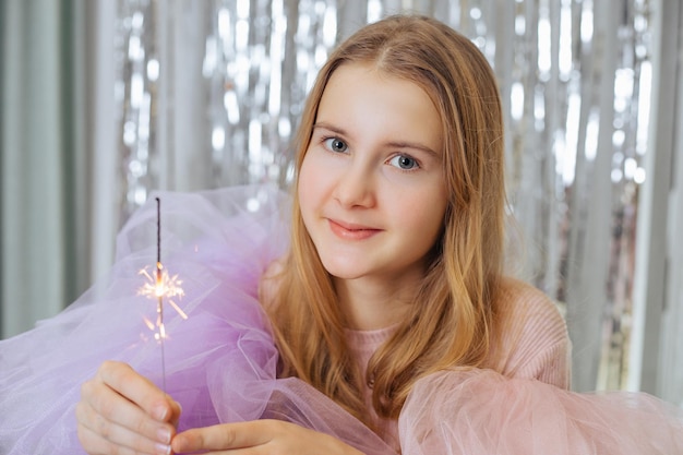 Charmantes, blondes, lächelndes Mädchen mit festlichem lila und weiß-rosa Kleid, das brennendes bengalisches Licht drinnen hält, möglicherweise in einem Café mit dekoriertem Hintergrund aus silbrigem Flitter