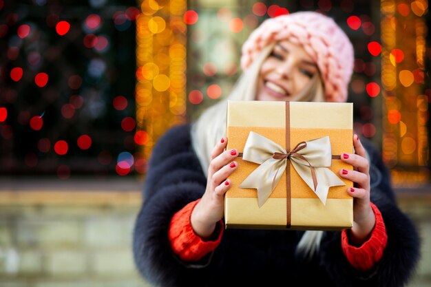 Charmantes blondes Mädchen mit Pelzmantel und Mütze zieht eine Geschenkbox zu Ihnen. Platz für Text