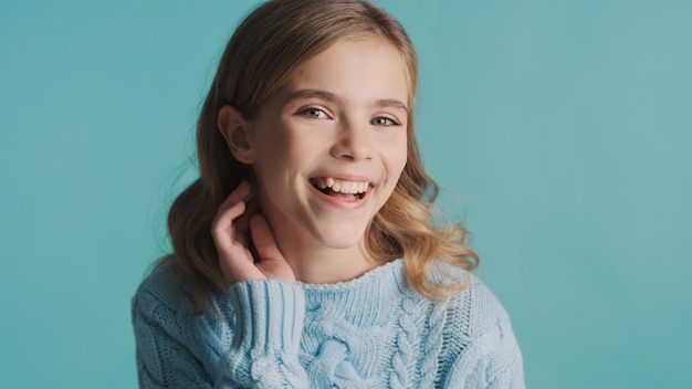 Charmantes blondes Teenager-Mädchen mit welligem Haar, das aufrichtig lächelt und vor der Kamera über blauem Hintergrund glücklich aussieht Glücklicher Ausdruck
