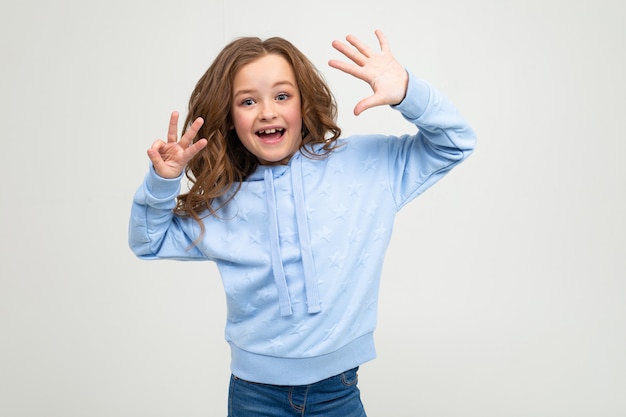 Charmantes jugendlich Mädchen in einem blauen Kapuzenpulli zeigt acht Finger auf einem hellgrauen Hintergrund