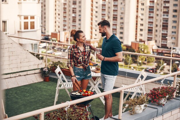 Charmantes junges Paar in Freizeitkleidung, das Grill zubereitet und lächelt, während es auf der Dachterrasse im Freien steht?