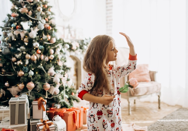 Charmantes kleines Mädchen im Pyjama hält eine Blume im hellen gemütlichen Raum mit dem Neujahrsbaum.