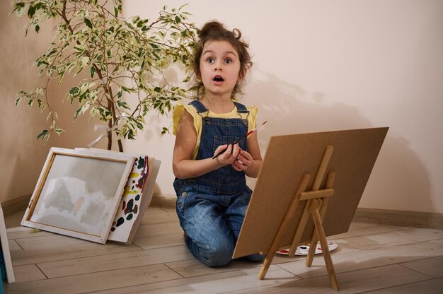 Charmantes kleines Mädchen in gelbem T-Shirt und blauem Denim, das einen Pinsel hält und mit Aquarell- oder Ölfarben auf Leinwand malt, die auf dem Boden neben gemalten Bildern zu Hause sitzen Kunstkonzept für Kinder