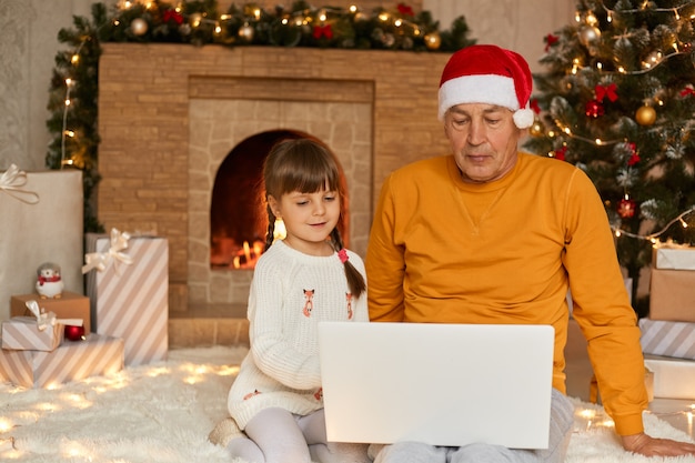 Charmantes kleines Mädchen mit Großvater sitzend und unter Verwendung des digitalen Laptops während Heiligabend sitzend auf Boden auf weichem Carper nahe Tannenbaum und Kamin, Familie, die auf Bildschirm konzentriert konzentriert.