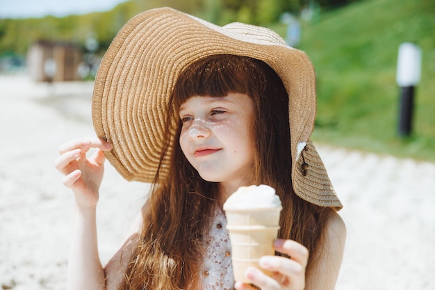 Charmantes kleines Mädchen mit Hut isst Eis am Strand Sommerurlaubskonzept