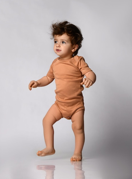 Charmantes lockiges Baby in beigem Körper macht seine ersten Schritte auf grauem Hintergrund