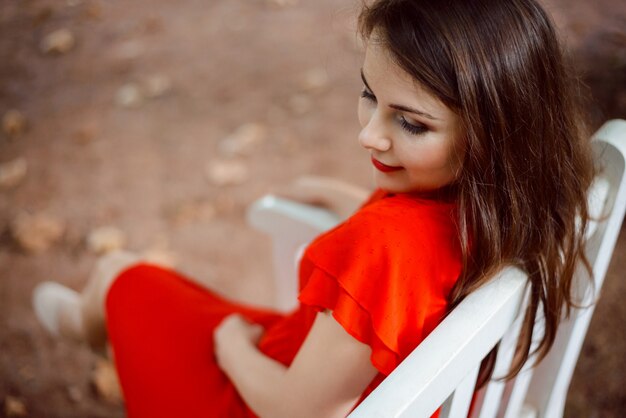 Charmantes Mädchen im bunten roten Kleid, das auf Bank im Park sitzt und auf jemanden wartet