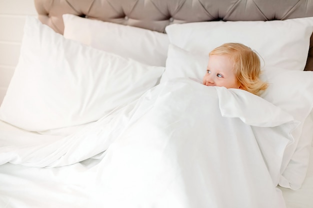 charmantes Mädchen liegt in einem weißen Bett unter einer Decke zu Hause und schaut weg