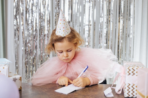 Charmantes Mädchen von 5 Jahren mit pinkfarbenem Festivalkleid, Partyhut, Sternendekoration auf ihrem Gesicht, Geschenkboxen, Schreiben von Wunschkarten oder Briefen im Café mit dekoriertem Hintergrund aus silbernem Flitter