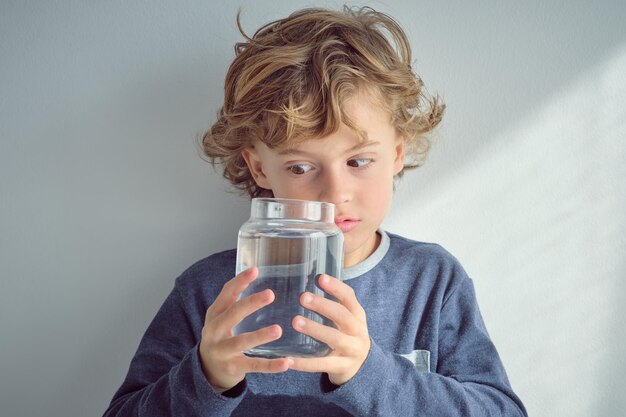 Charmantes, neugieriges Kind mit lockigen blonden Haaren im warmen Pullover, das in eine Glasflasche mit transparentem, klarem Wasser schaut, während es vor einer weißen Wand steht