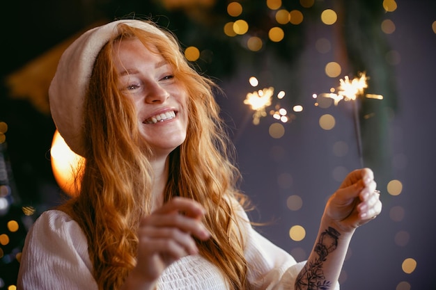 Charmantes rothaariges Mädchen in einer Weihnachtsmütze mit Wunderkerzen in den Händen, die in die Kamera lächeln.