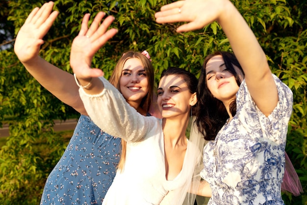 Foto charming joven novia en un vestido blanco y dos damas de honor con velos rosados posando bloqueando sus caras del sol con sus palmas alegre fiesta de soltera en el parque de verano retrato en primer plano