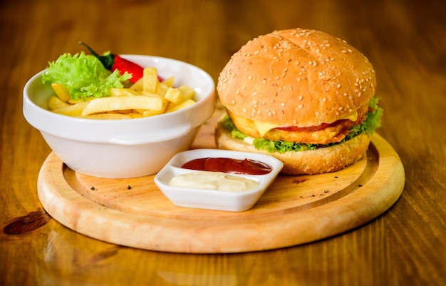 Cheat meal Hambúrguer delicioso com sementes de gergelim Menu de hambúrguer Lanche de alto teor calórico Hambúrguer e batatas fritas e molho de tomate na tábua de madeira Conceito de fast food Hambúrguer com queijo, carne e salada